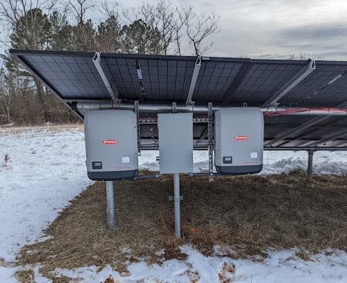 Two string inverters and an isolator box mounted beneath a ground-mounted solar array in snow