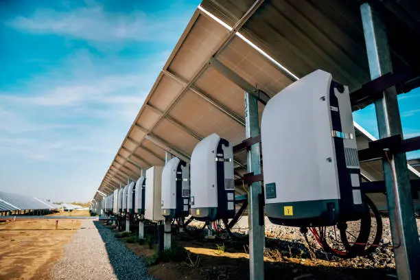 A long row of white string inverters mounted under a large ground-mounted solar array